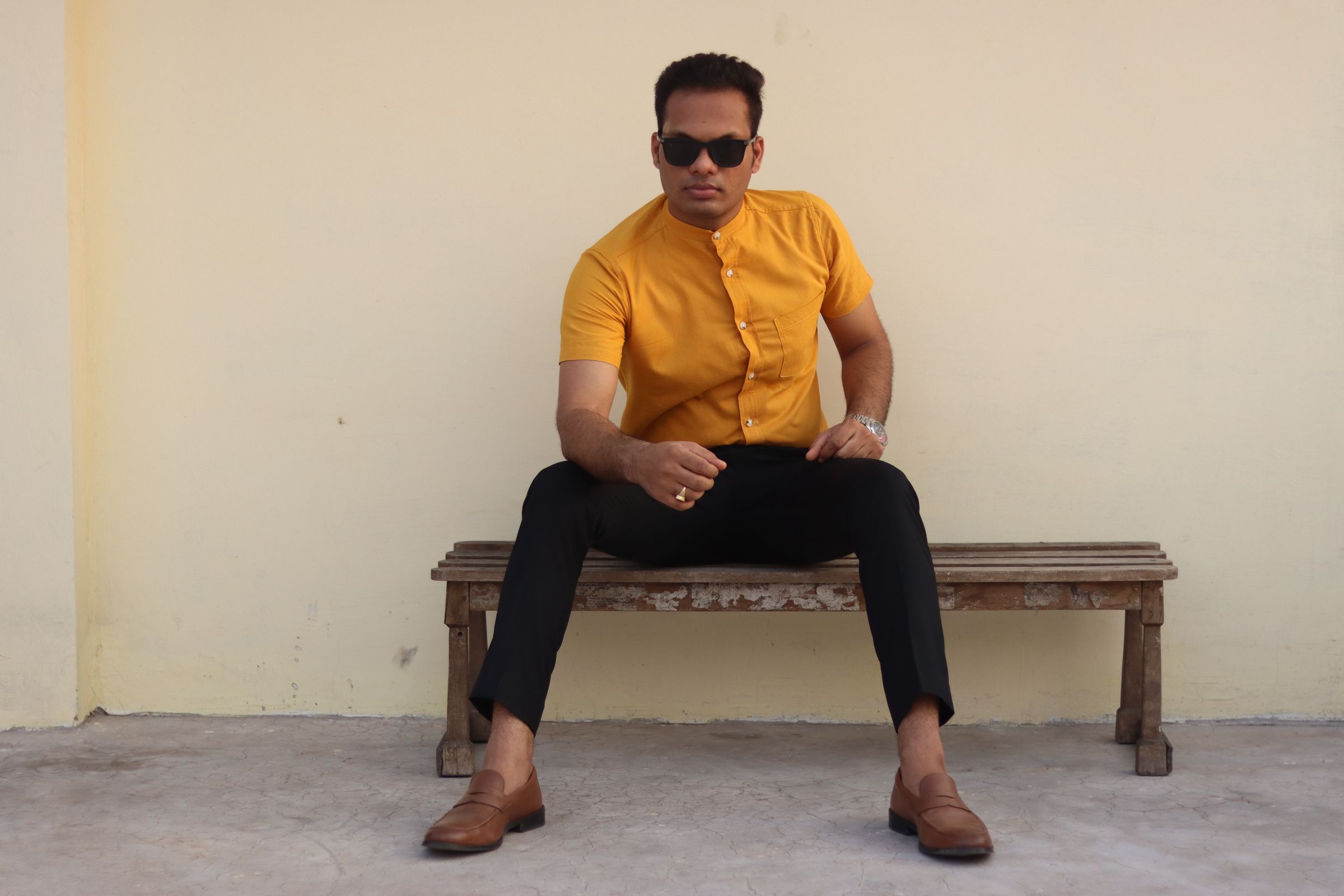Darkhill — Landscape: mustard band-collar shirt + black trousers on rustic bench against wa