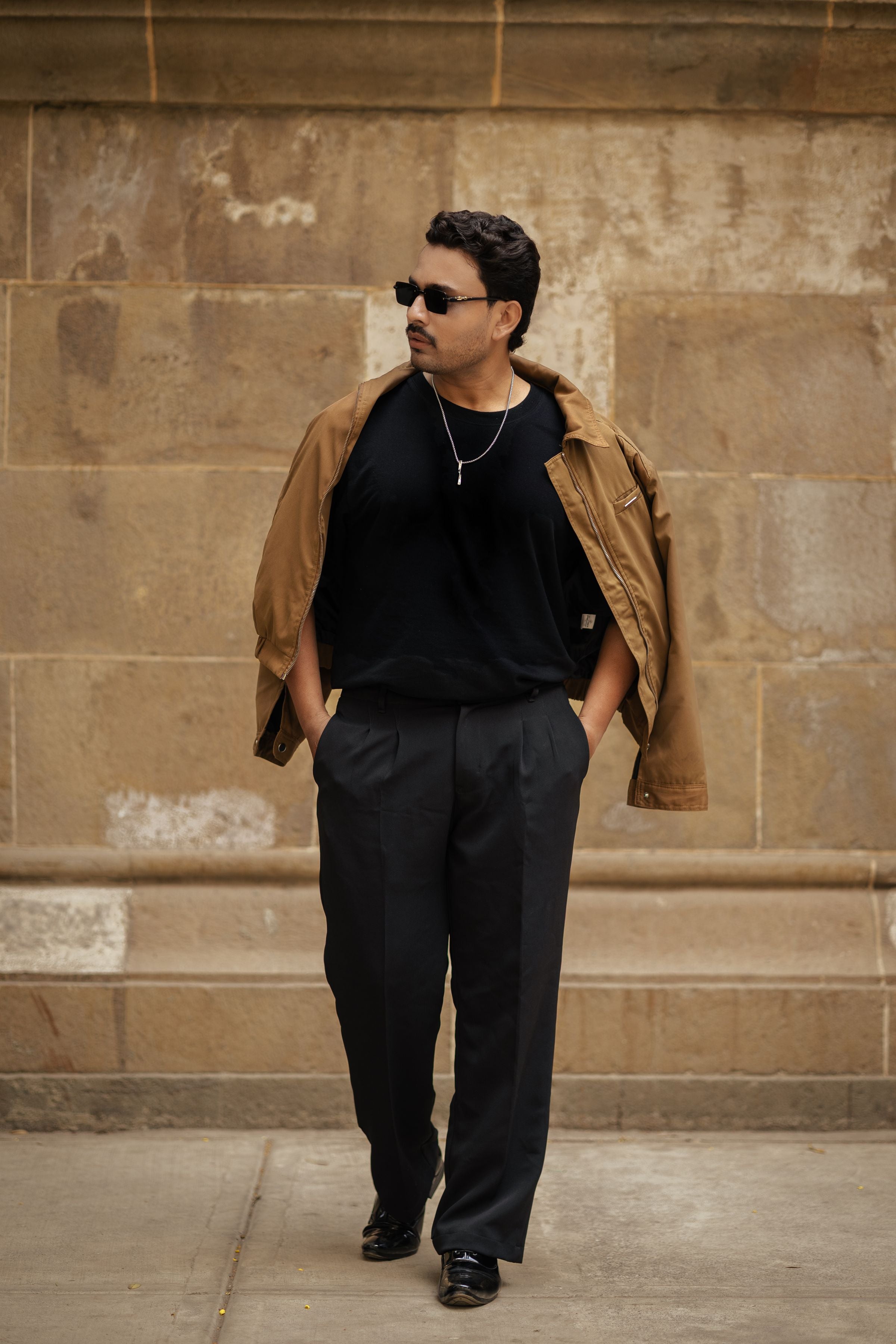 Darkhill — Camel overshirt + black tee + black trousers by stone architecture, sunglasses