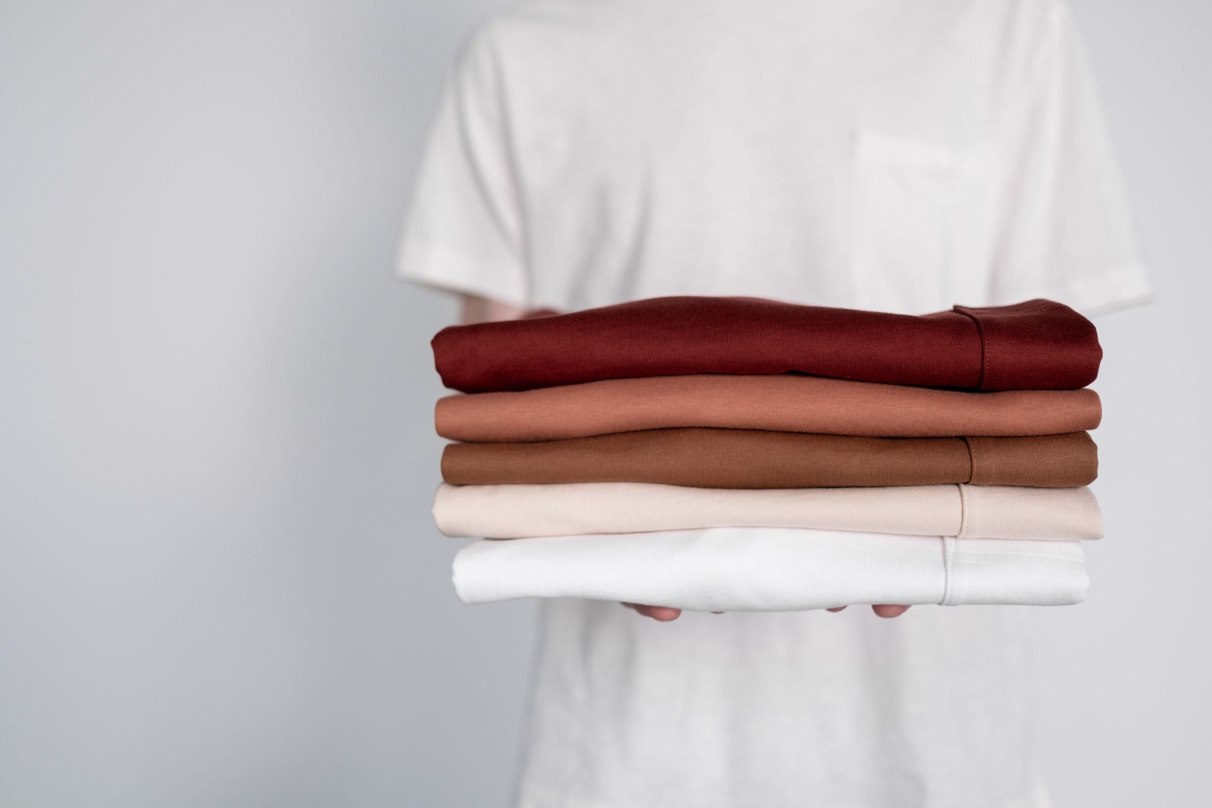 Darkhill — Hands holding stack of folded shirts in earthy wine/tan/cream tones on soft grey